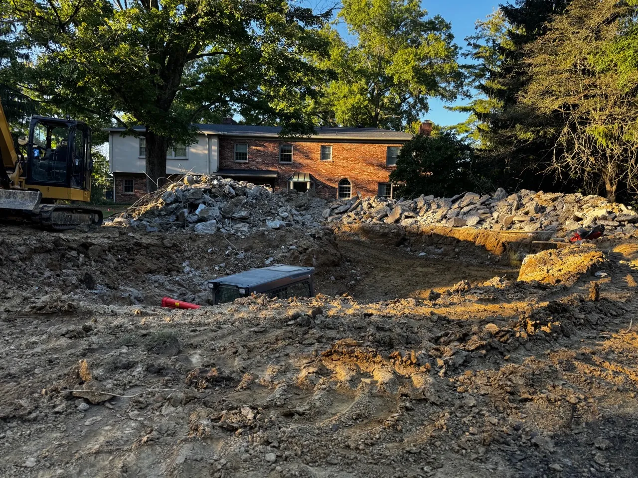 Rubble pile and open hole after a pool shell was broken out, with an excavator at the edge and a brick house in the background.